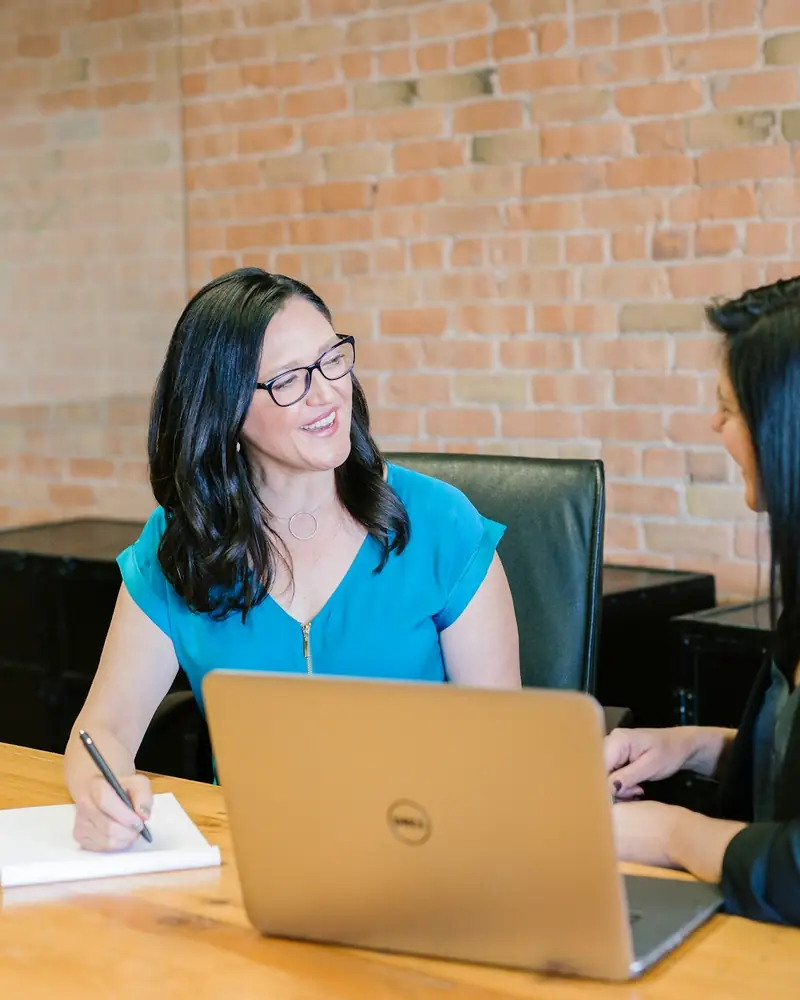 Two people talking across a table with a laptop and a notebook